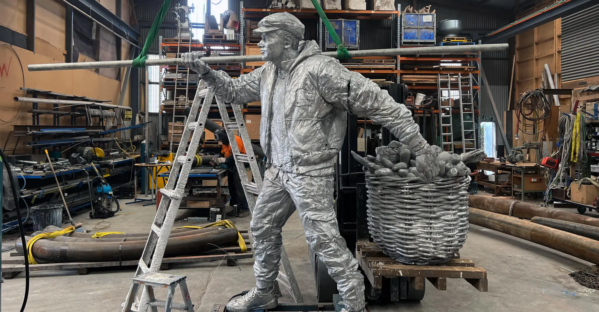 Silver sculpture of a vegetable hawker with a basket full of produce, positioned in a workshop surrounded by tools and equipment.