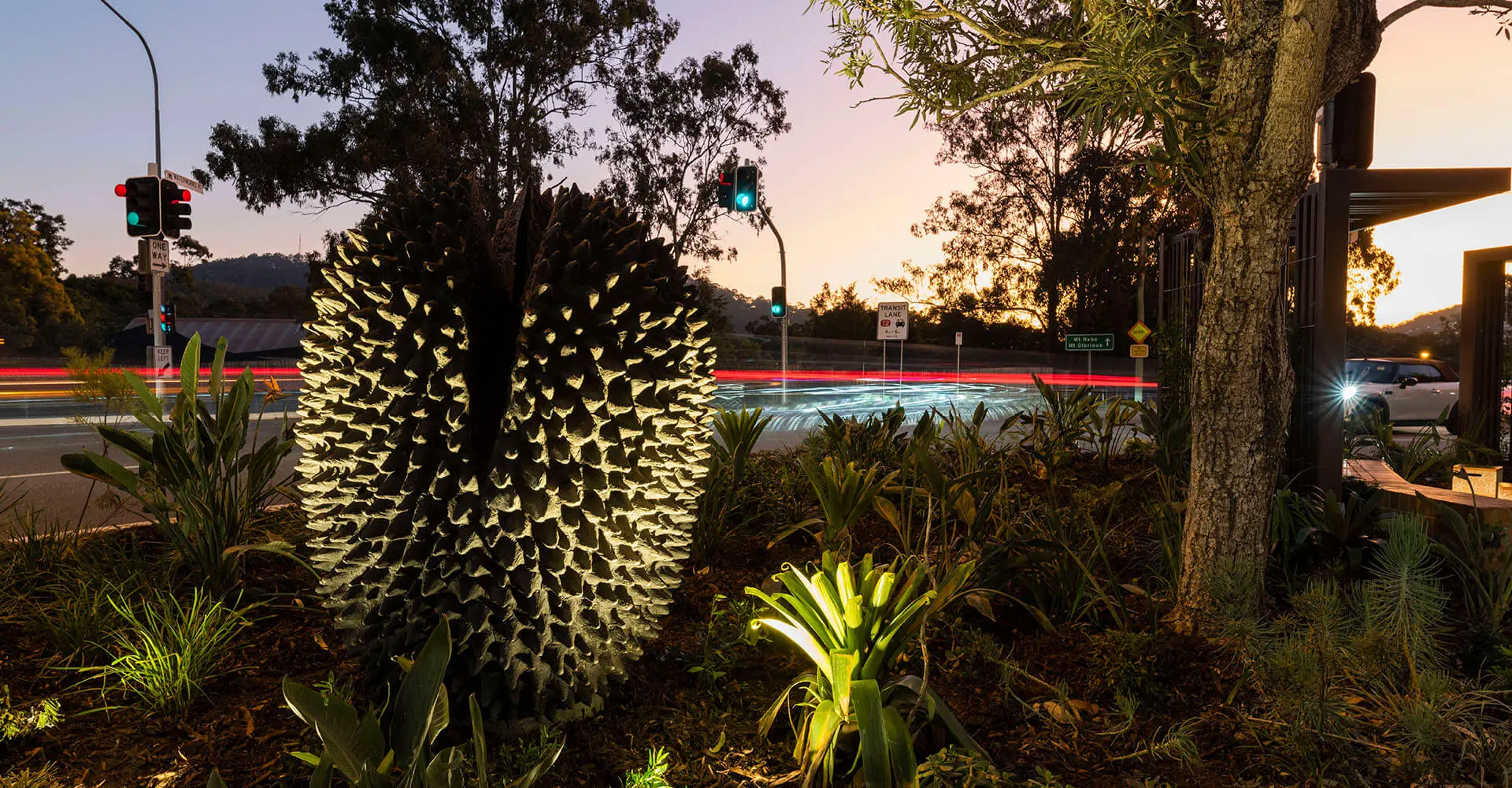Twilight scene at an intersection showcasing a luminescent, pinecone-like sculpture surrounded by verdant plants. The ambient light captures the moving cars' streaks and the setting sun, titled 'Crowsash Pods'.