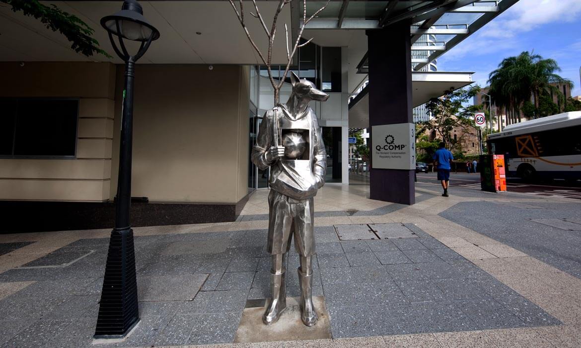 A metallic sculpture, 'The Guardian',  of a humanoid figure with fox-like facial features stands on a city sidewalk. The sculpture holds a thin, bare tree and has a sphere embedded in its torso. Adjacent to it stands a street lamp, with a building's facade in the background. In the distance, a pedestrian walks by, and a bus is visible near a sign that reads 'Q-COMP'.